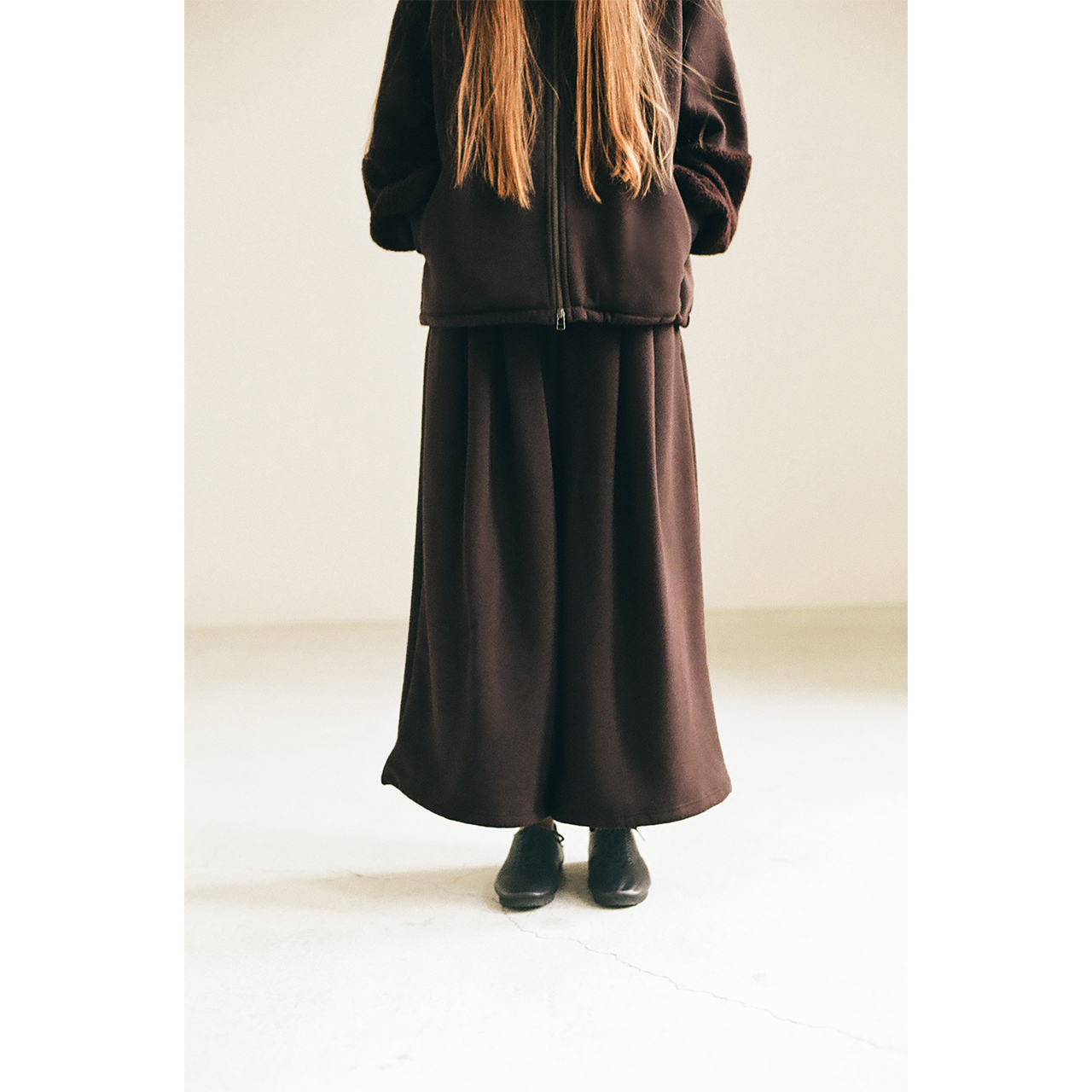 Back view of a model wearing a dark brown zip blouson in the Mochi 2026 Autumn Winter image look.