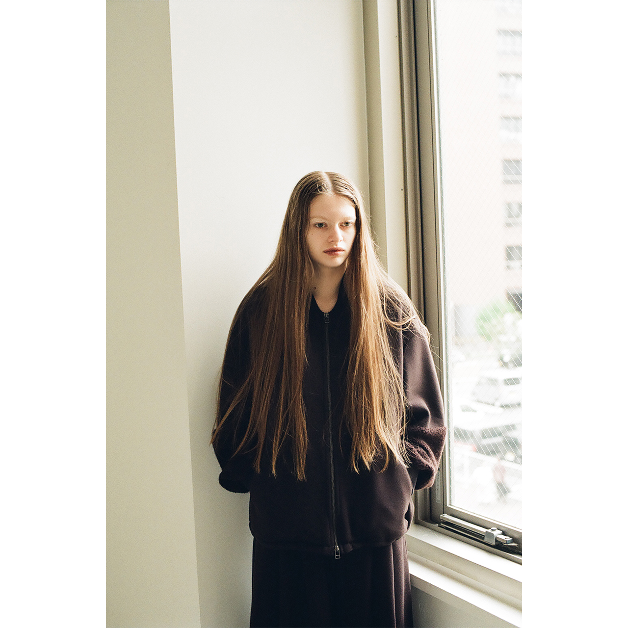Model wearing a dark brown zip blouson by the window in the Mochi 2026 Autumn Winter image look.